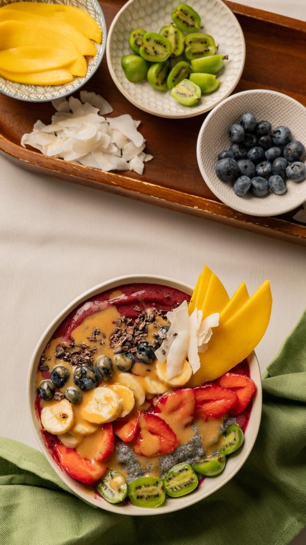 Açaí Bowl mit Schälchen voller Mango, Mini-Kiwi, Blaubeeren und Kokoschips auf einem Holztablett