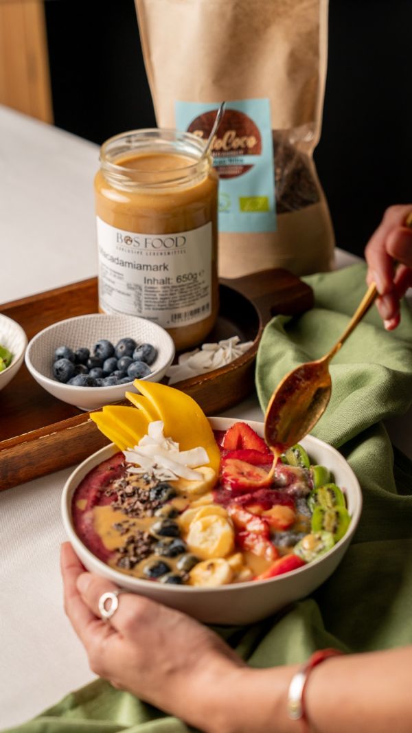 BOS FOOD Macadamiamark im 650-Gramm-Glas, daneben wird Nussmus über eine Açaí Bowl gezogen